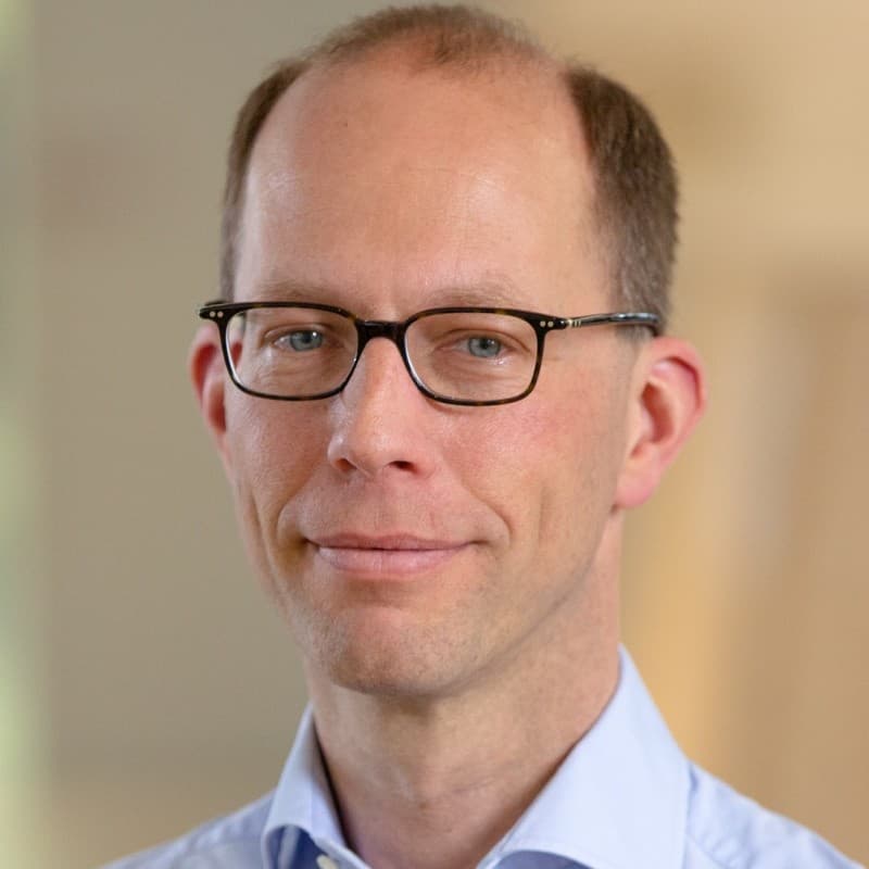 Smiling man with glasses, blue eyes, and a receding hairline wearing a light blue collared shirt.