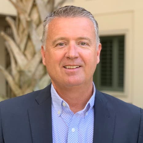 Smiling middle-aged man with short gray hair wearing a dark blazer and patterned shirt.