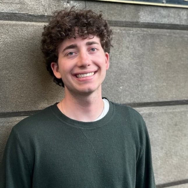 Smiling young man with curly brown hair wearing a dark green sweater against a wall.