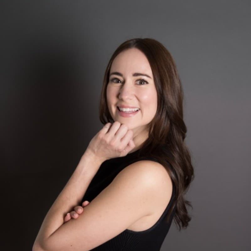 Smiling woman with long brown hair, hand under chin, posing against a solid gray background.