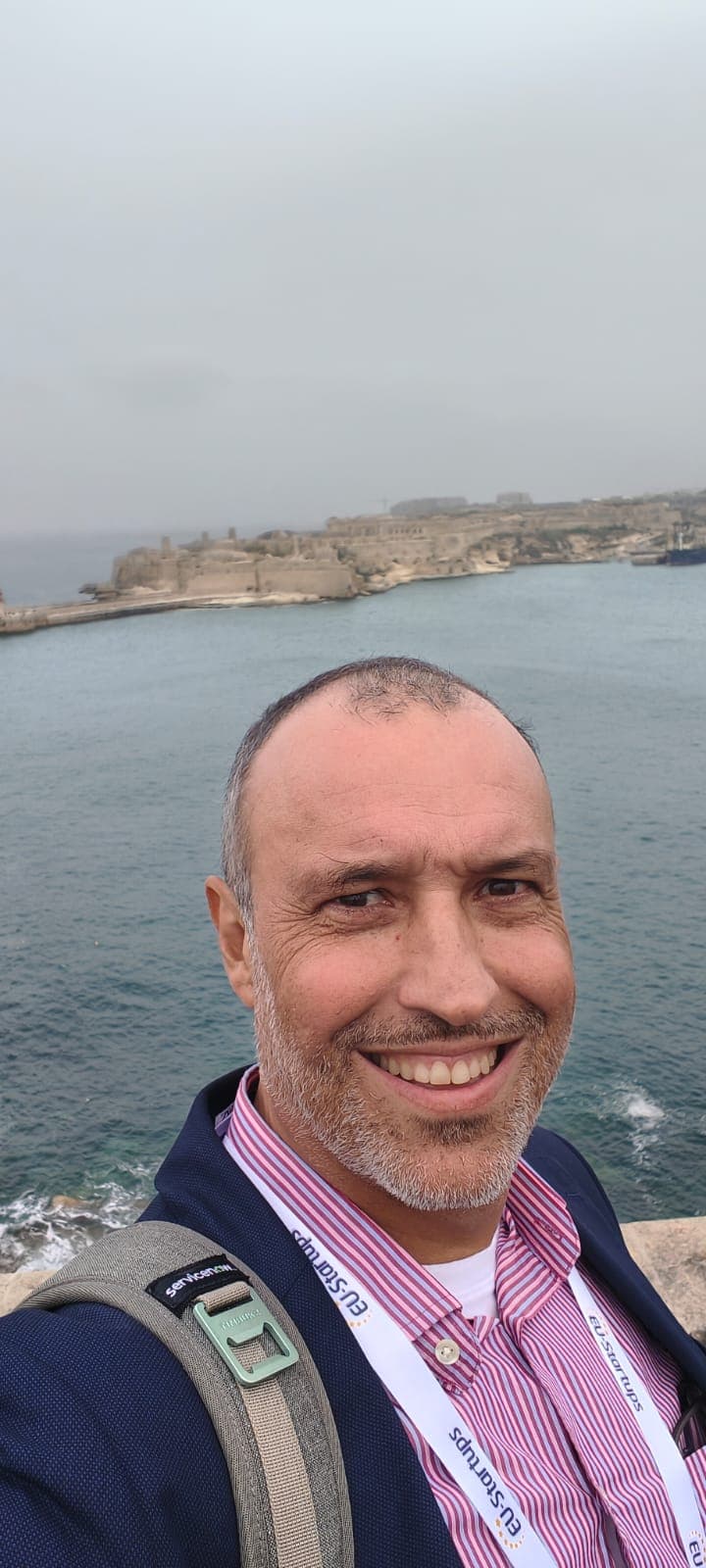 Smiling man wearing an EU-Startups lanyard with a scenic coastal fortress in the background.
