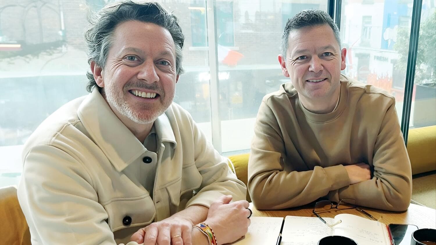 Two men smiling at a table with a notebook in front of a bright window.
