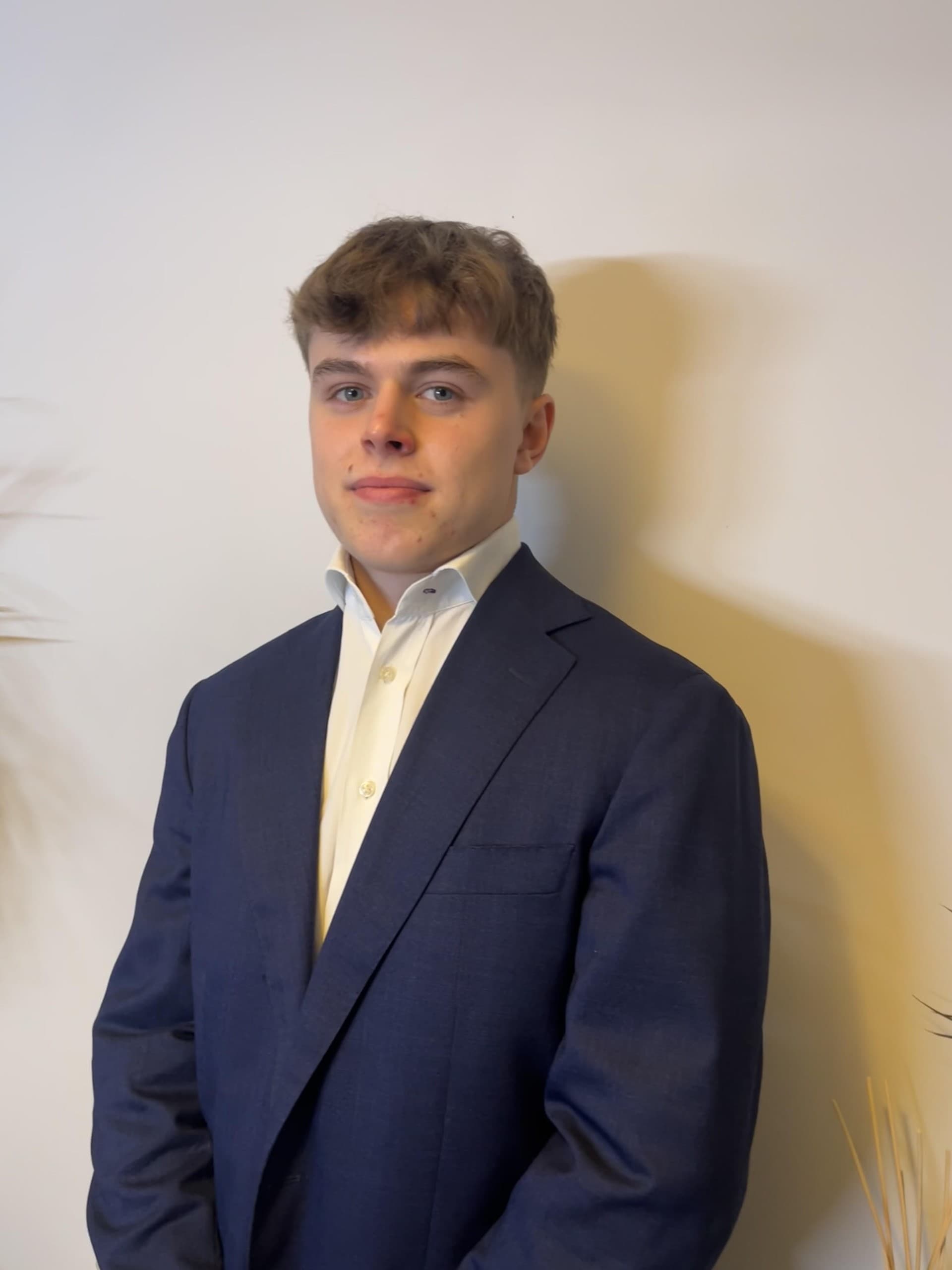 Young man with brown hair in a navy blue suit jacket and white collared shirt.